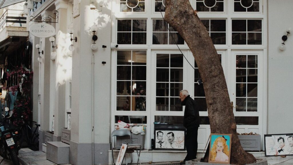 An elderly artist showcases portraits on a city street under a tree.