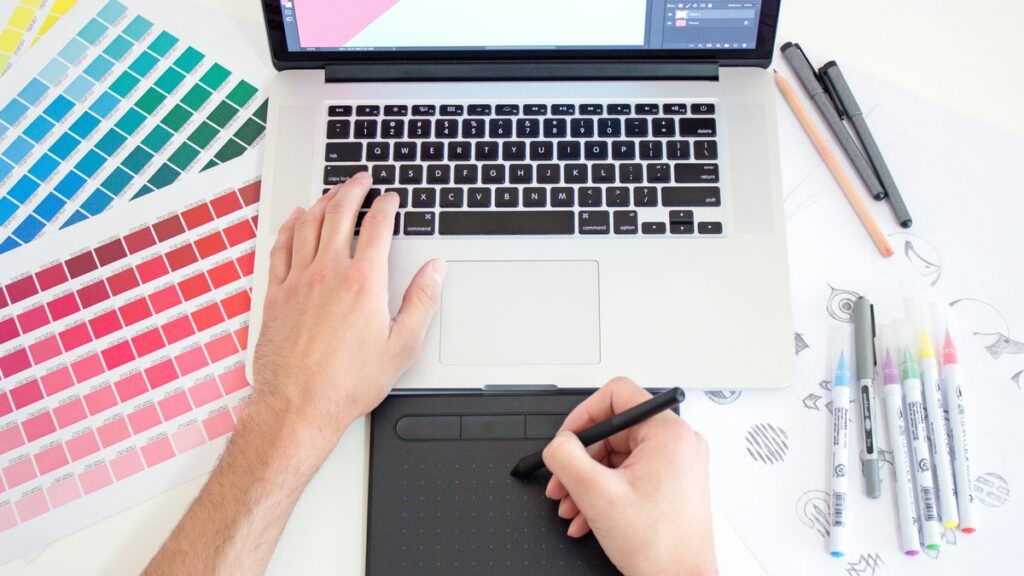 A person using a digital drawing tablet and laptop with color swatches and markers